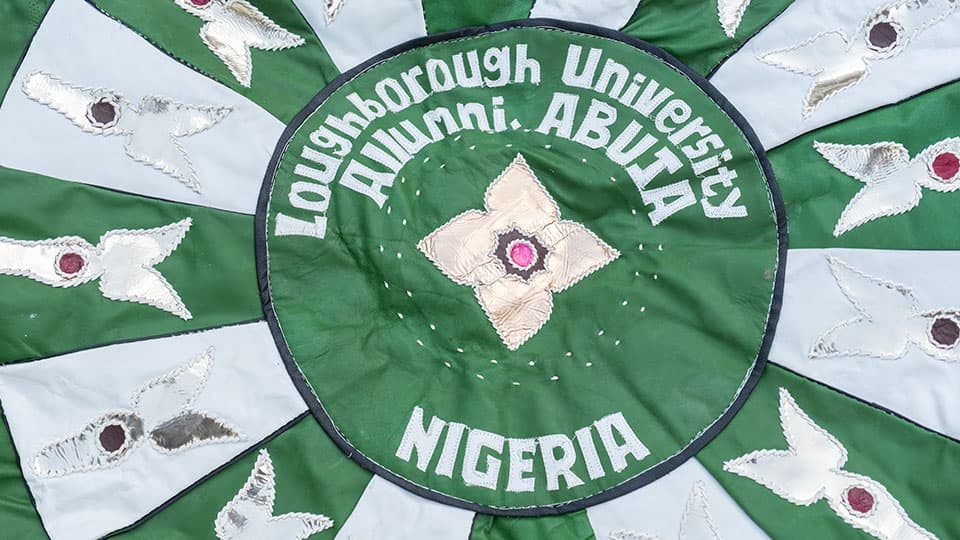 A green, white and gold handmade tapestry which has the words Loughborough University Alumni Abuja, Nigeria sewn in white text on top of it.